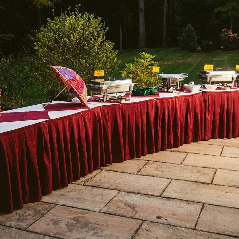 Outdoor buffet table with red skirting, chafing dishes, and colorful decorative umbrellas on a patio.