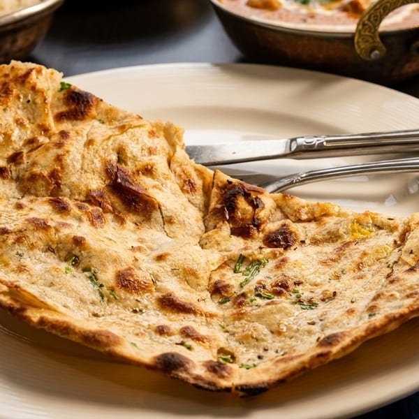 A golden-brown naan bread with herbs on a white plate, with cutlery and other dishes in the background.